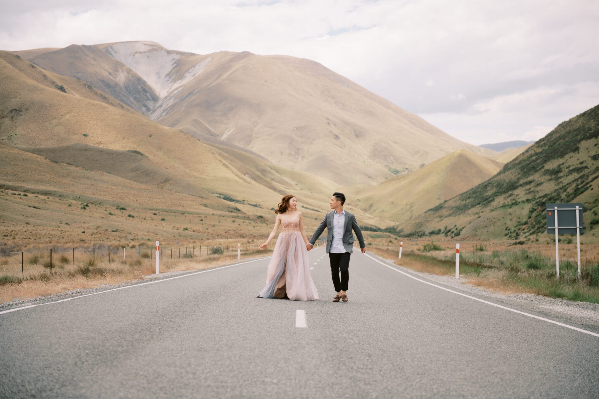 Queenstown Tekapo New Zealand Heli Wedding Elopement Pre-Wedding Shoot Photographer クイーンズタウン　テカポ　ニュージーランド　エロープメント 前撮り　フォトウェディング　結婚式 | A couple holding hands while walking down the center of a deserted road in Tekapo during their pre-wedding shoot, with mountains in the background.